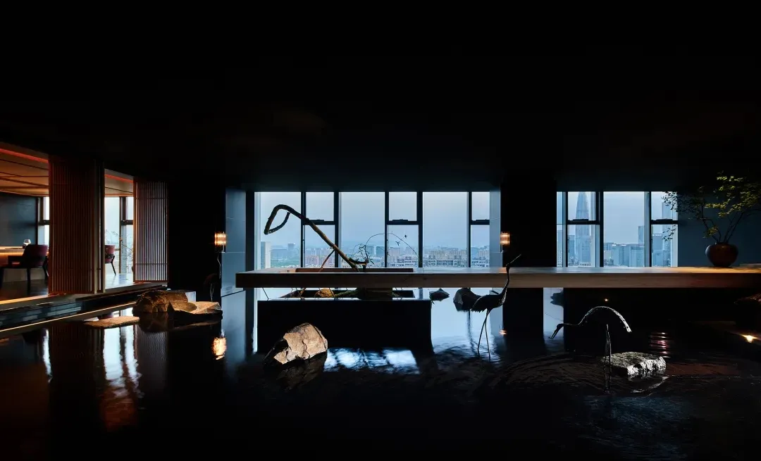 A minimalist hotel suite featuring a water feature with stones and a bird sculpture, overlooking a cityscape through large wi