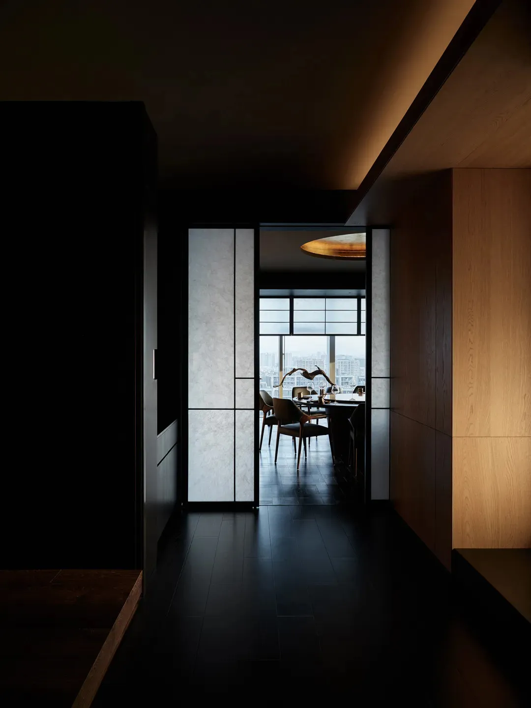 Modern minimalist hallway with wooden walls and a glass partition, leading to a dining area with a round ceiling light and a 