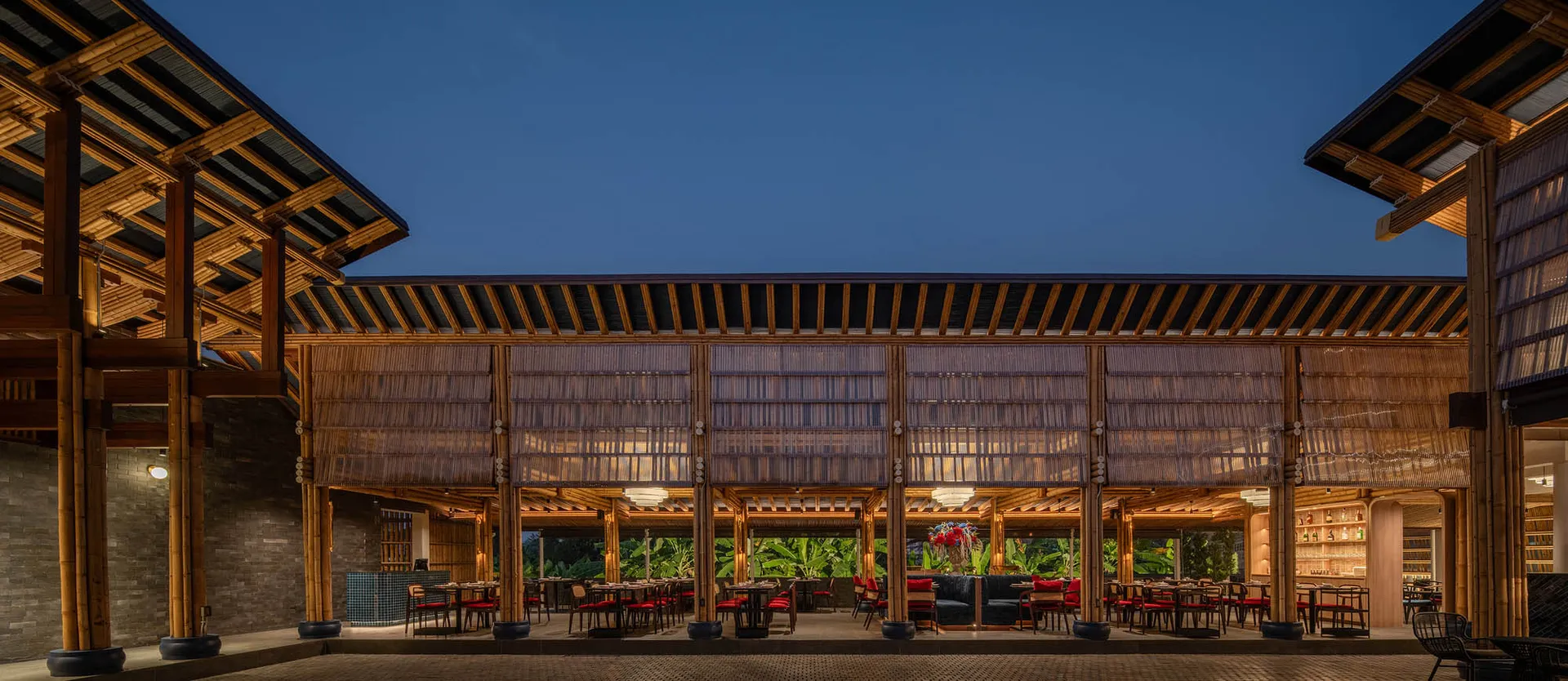 Outdoor area of a modern restaurant featuring bamboo and wooden structures, with tables and chairs arranged under a large roo