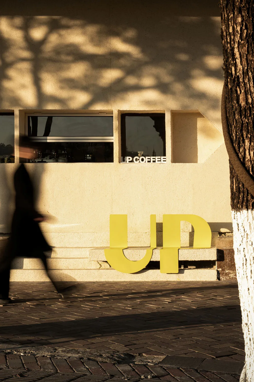 一間現代咖啡店外觀，黃色「UP COFFEE」招牌、混凝土牆面，以及樹影投射在立面上。