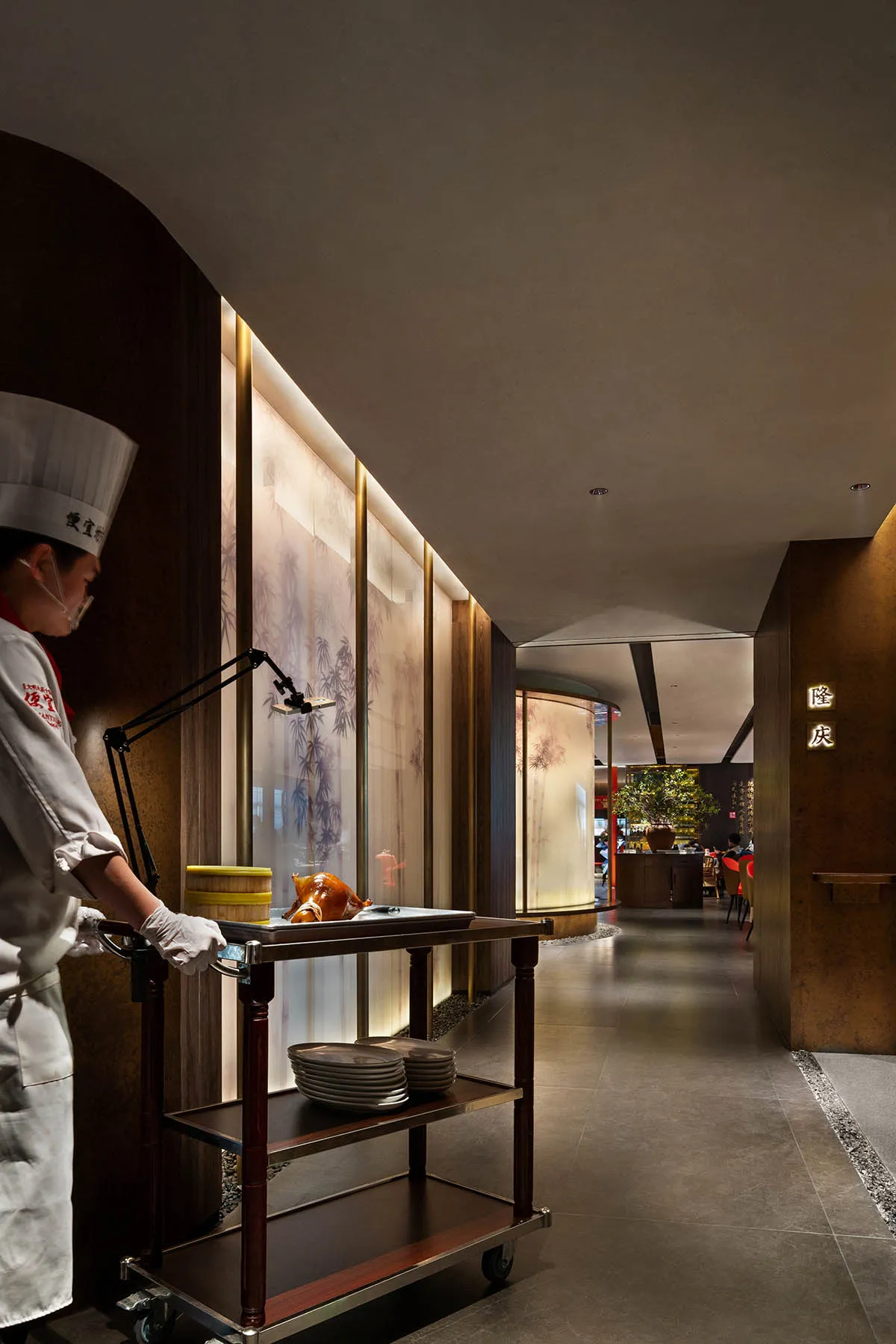 A chef pulls a cart with dishes in a modern restaurant interior made of wood, glass, and metal.