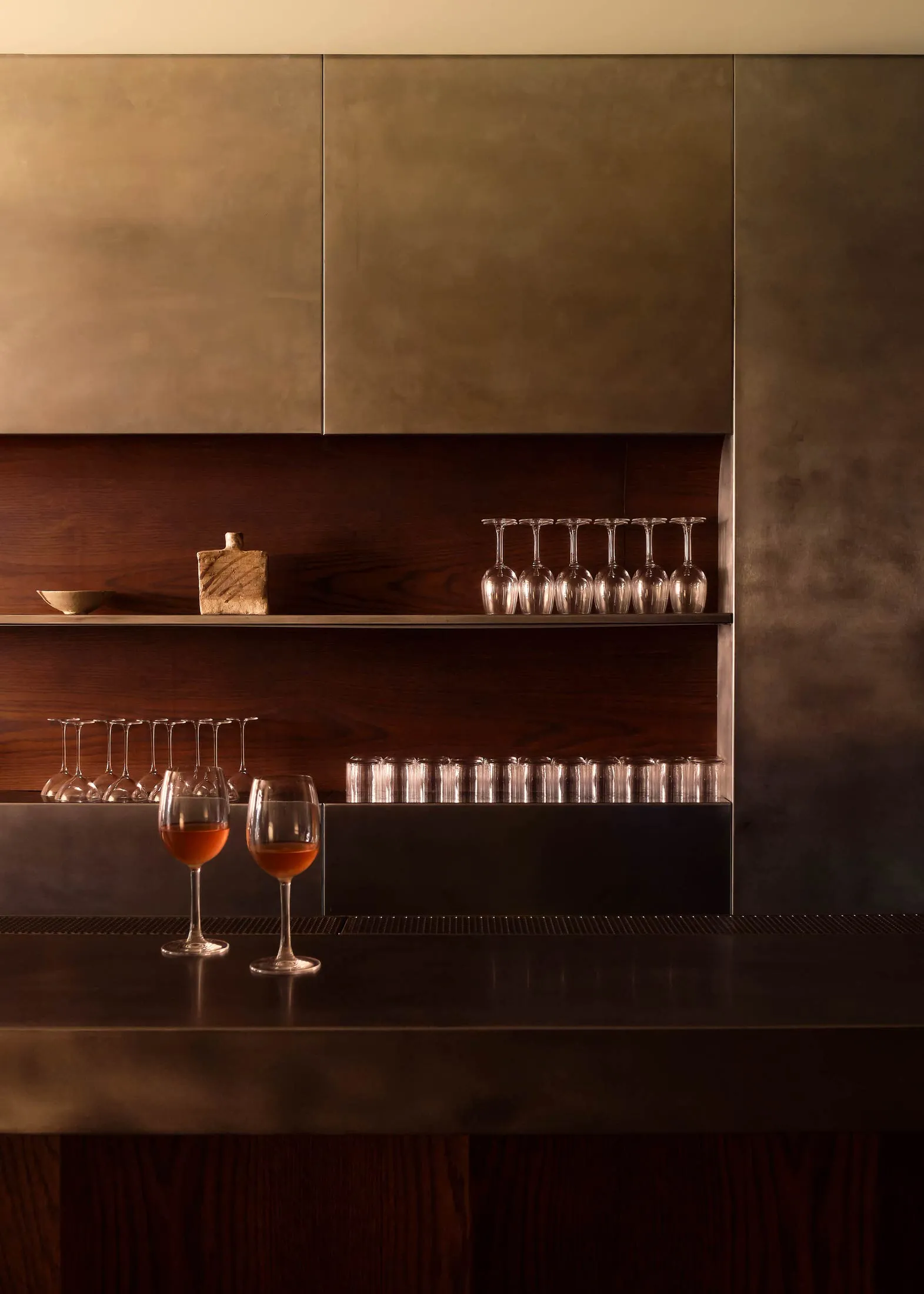 A minimalist bar counter with a metal finish, featuring a few drinks on the counter and an organized shelving unit with wine