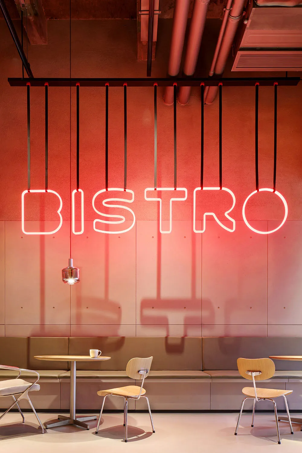 A minimalist industrial-style restaurant interior with exposed concrete walls, a red neon sign reading 'BISTRO', and simple s