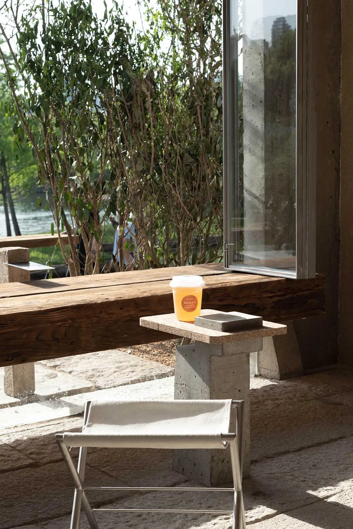 A modern coffee shop exterior with wooden seating and concrete structures, surrounded by greenery, with a cup of drink on a s