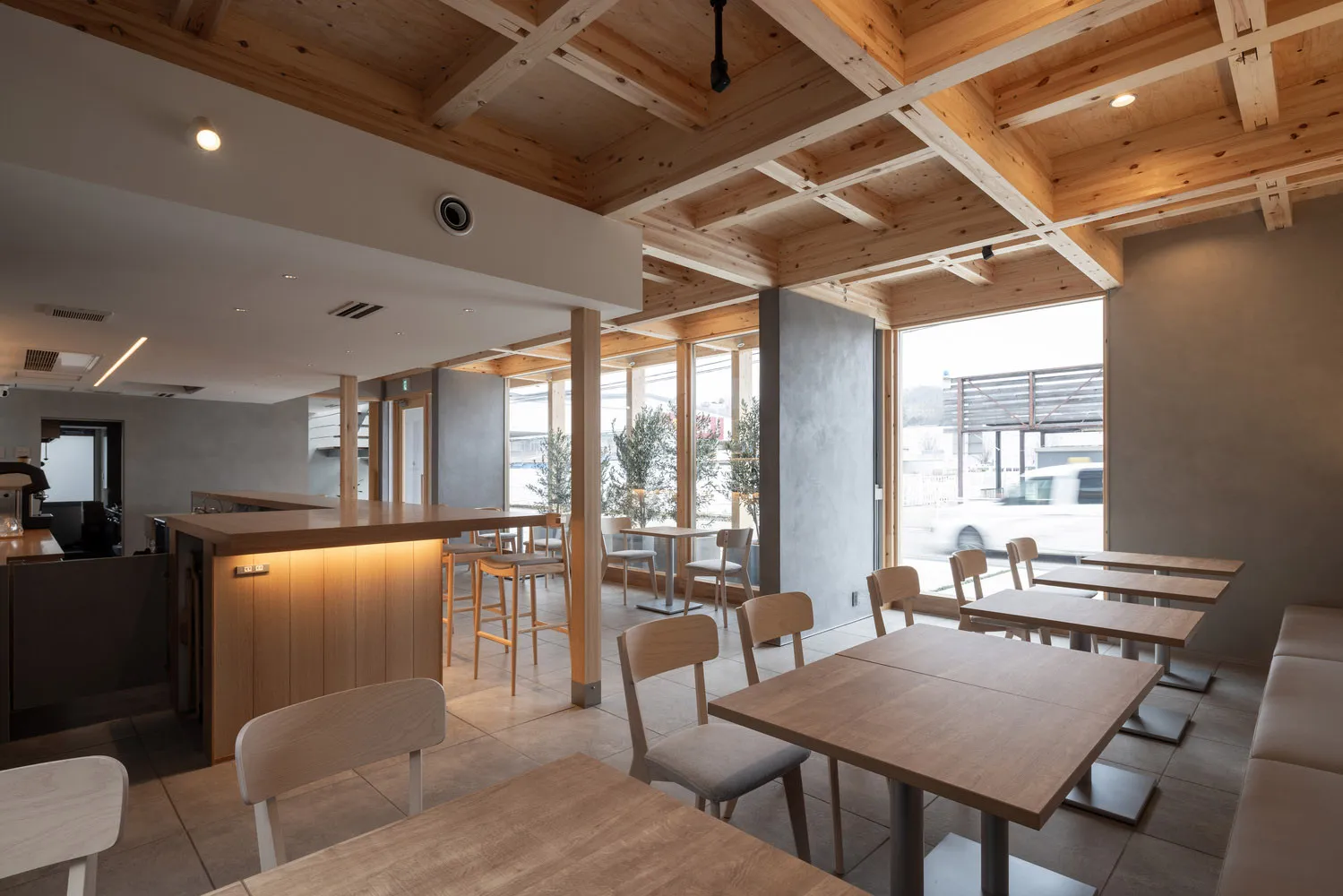 A well-lit, modern cafe interior with wooden tables and chairs, large windows, and exposed wooden beams on the ceiling.