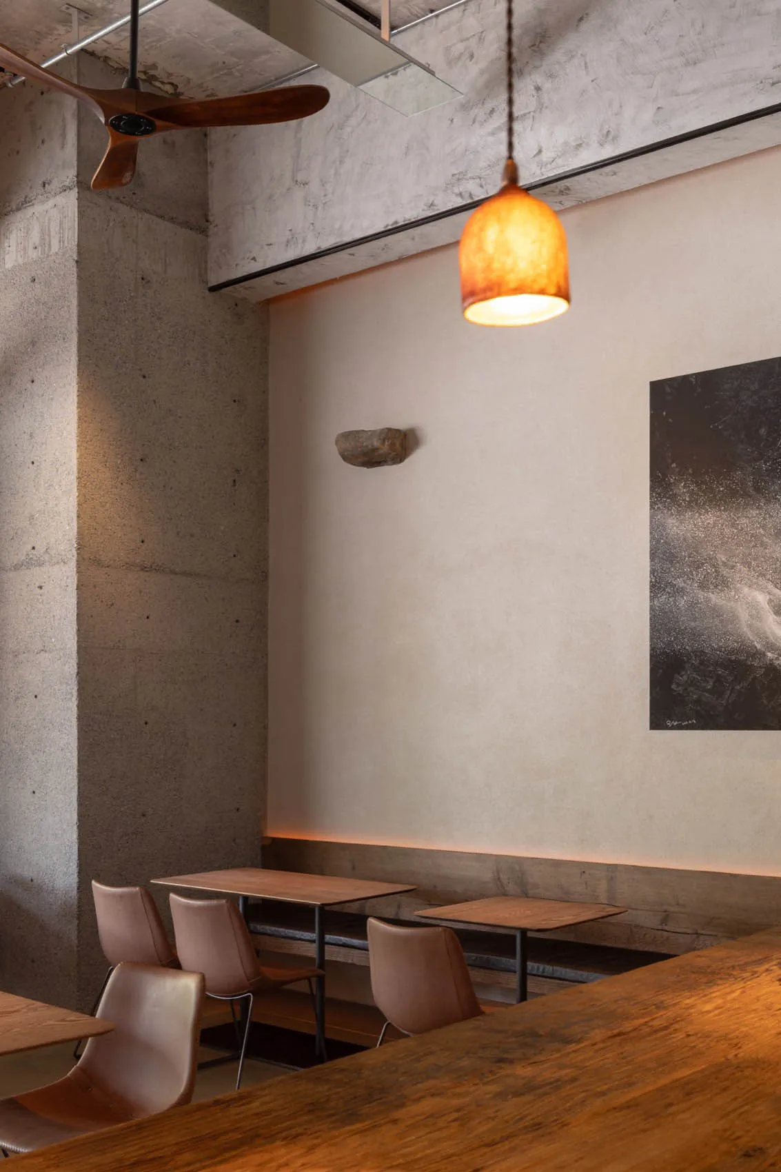A modern, industrial-style restaurant with concrete and wooden elements, featuring a wooden ceiling fan and warm pendant ligh