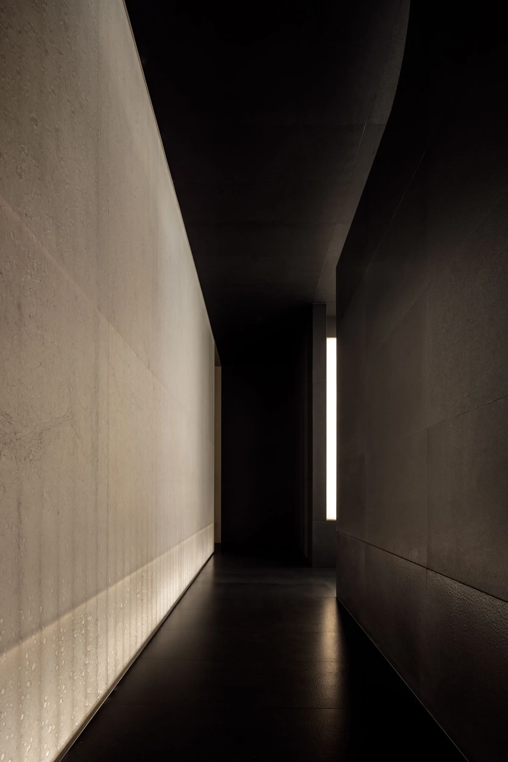 A minimalist corridor with concrete walls and floor, featuring a single illuminated window at the far end.