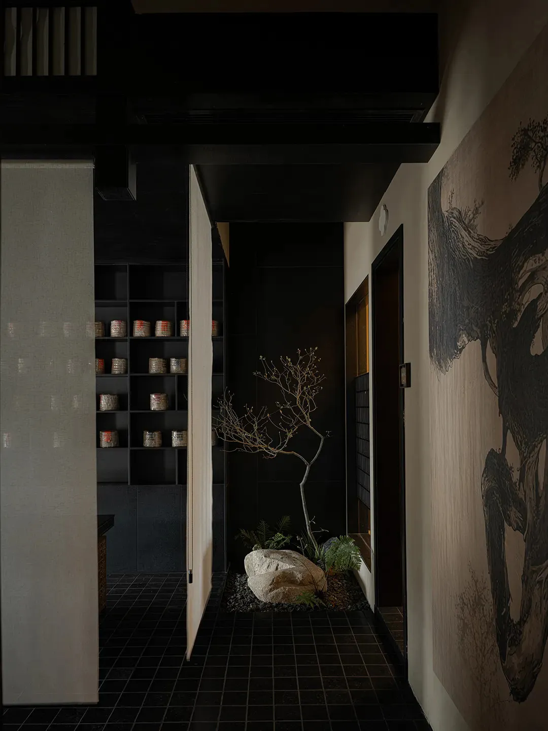 A modern entryway featuring dark-toned tiles, a small rock garden with bare trees, and a wall with large mural depicting tree