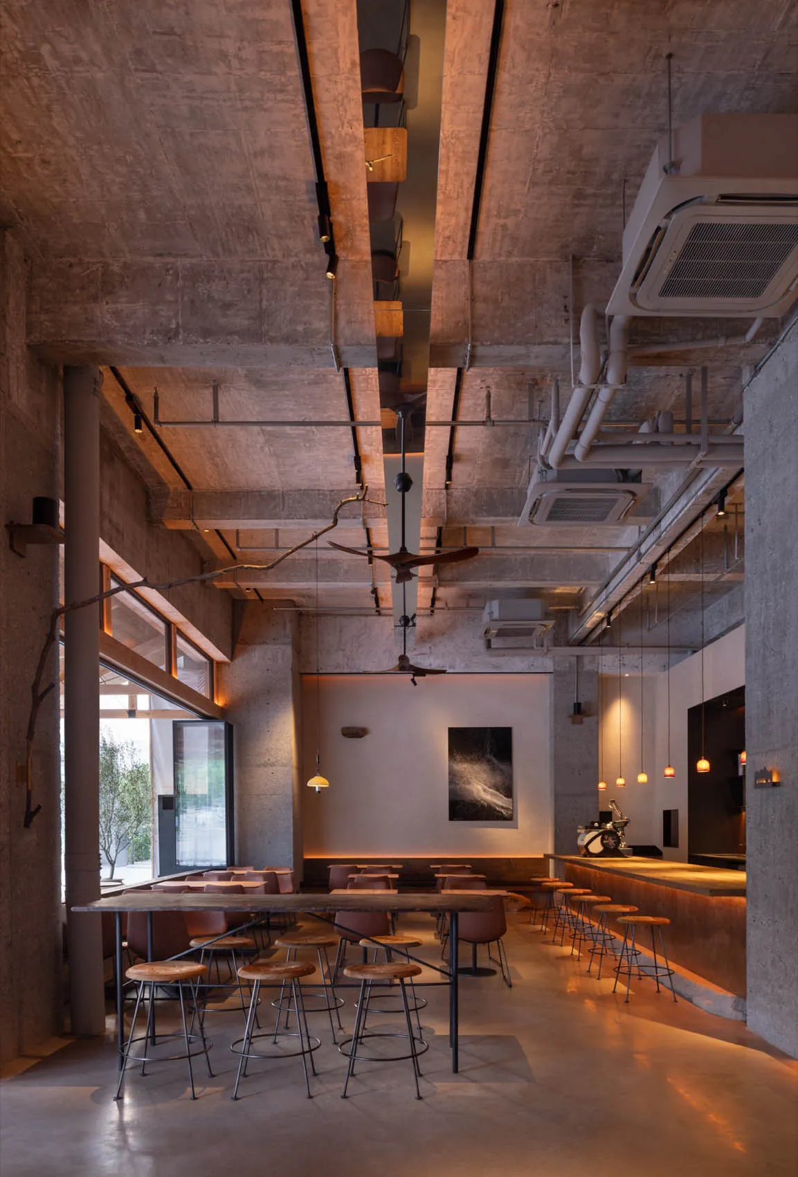 Industrial-style cafe interior with concrete walls, ceiling fans, and wooden stools.