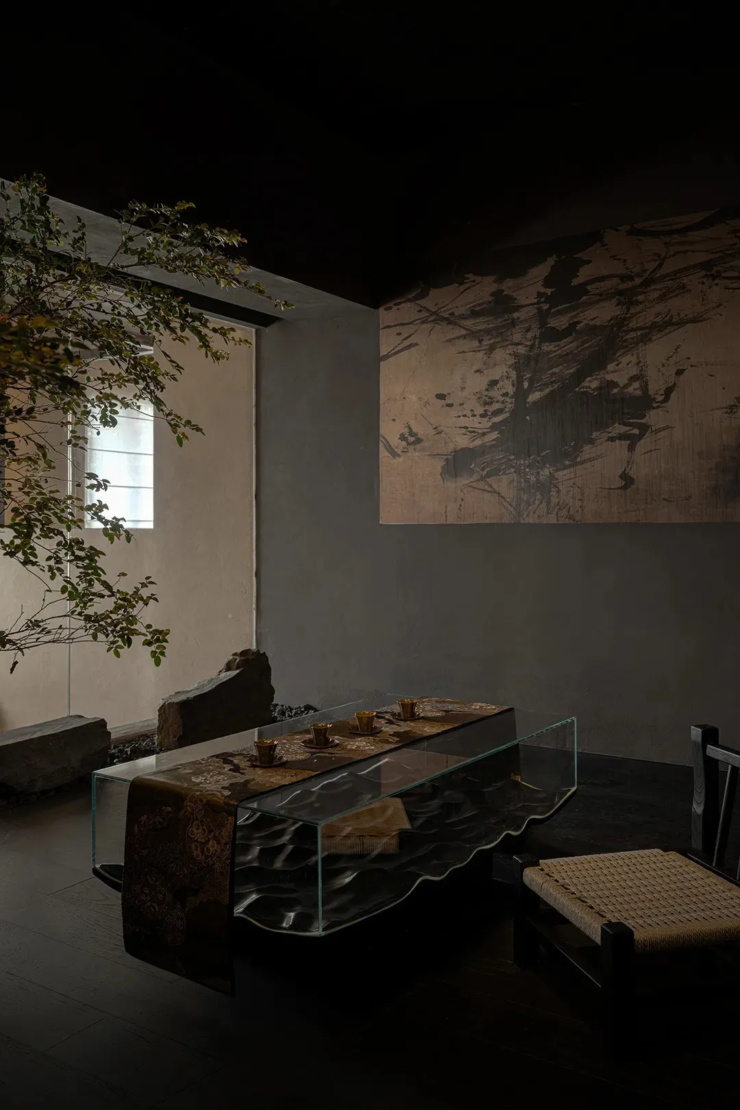 A minimalist tea room featuring a modern glass table with a dark, wavy base and a traditional Japanese hanging scroll hanging