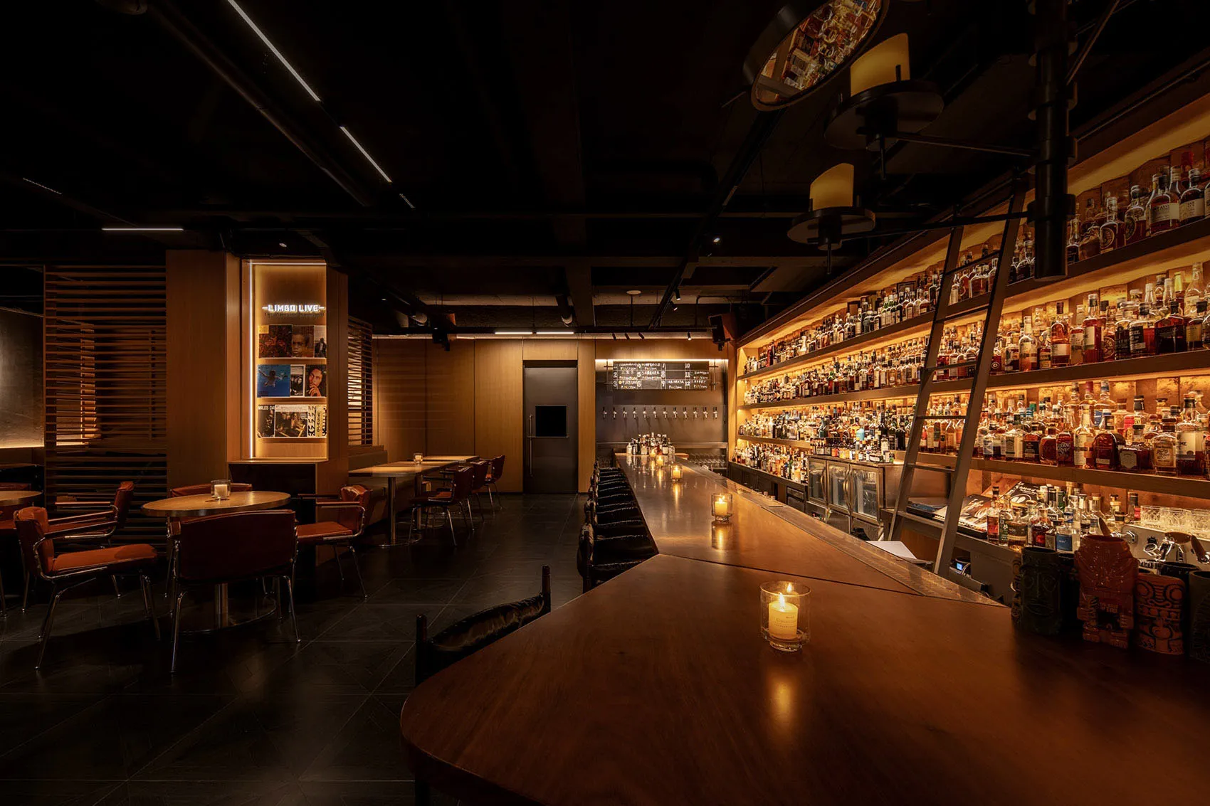 A dimly lit industrial-style bar with wooden counter, shelves lined with bottles, and a ladder.