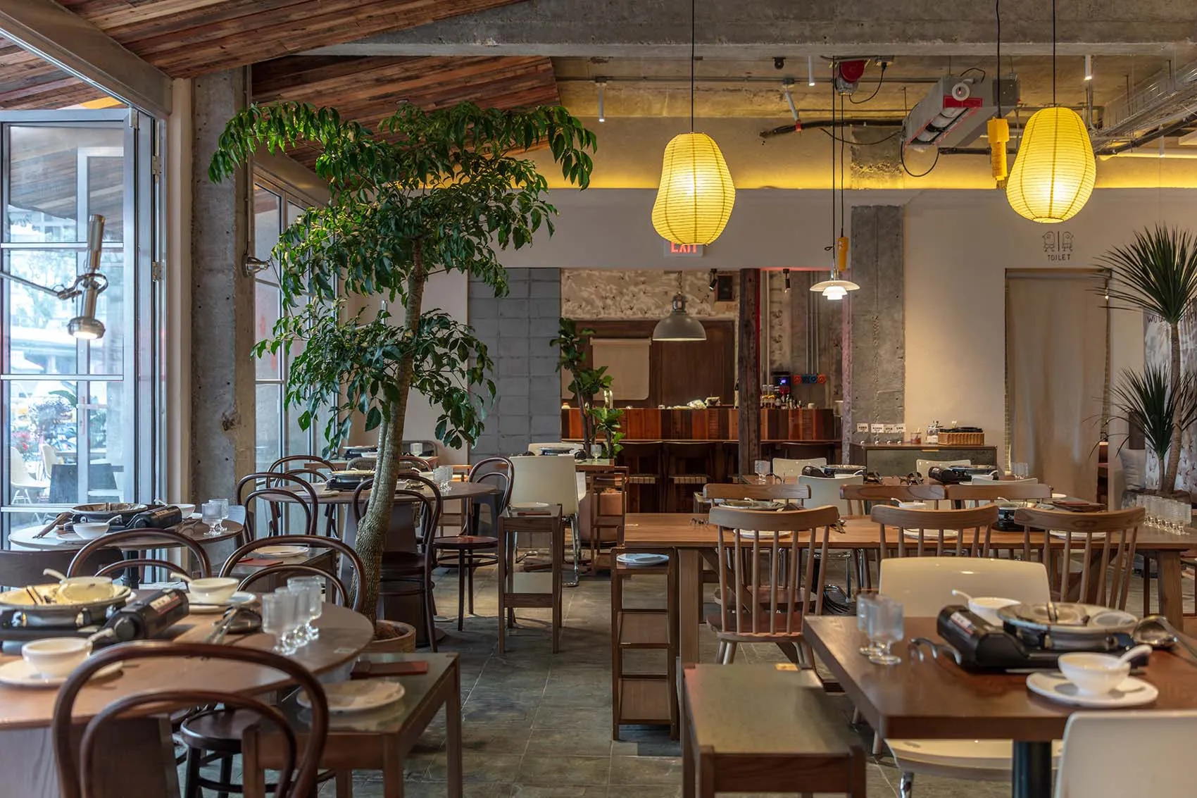 A spacious modern restaurant interior with wooden tables, concrete columns, and hanging lantern lights.
