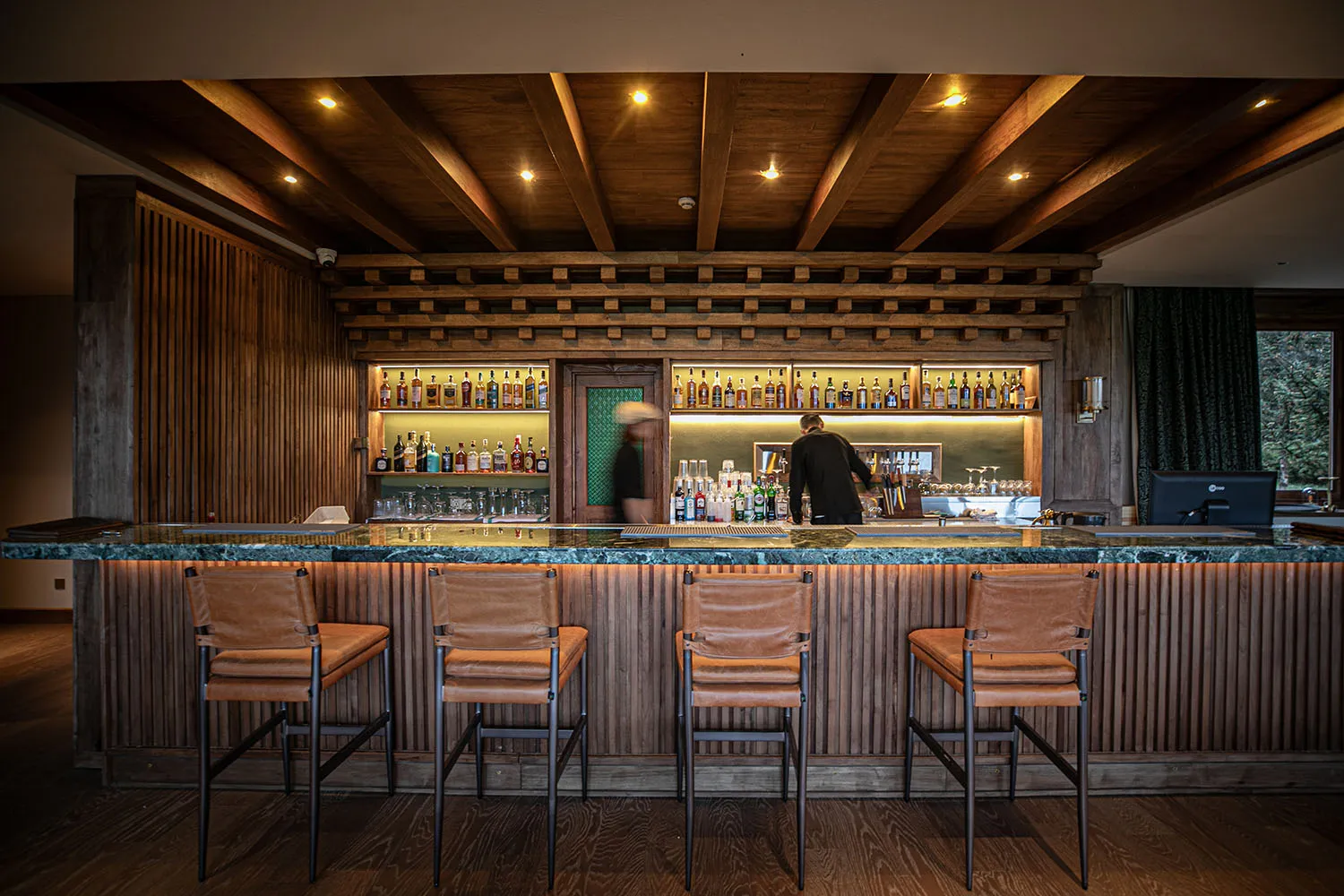 A modern bar with a wooden interior, featuring a counter, stools, and various bottles.