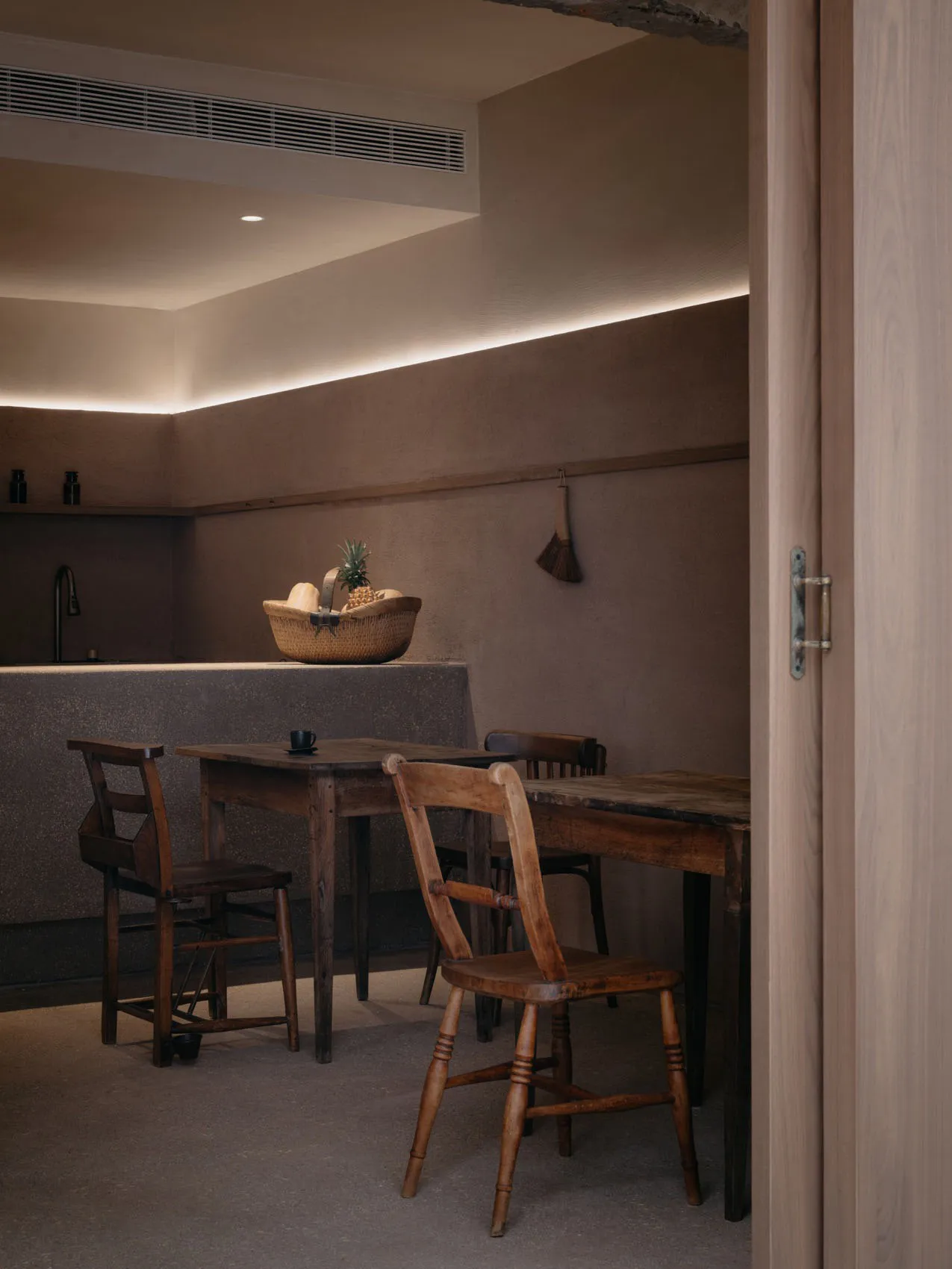 A minimalist residential kitchen with wood and concrete materials, featuring a basket of fruits and a vintage table and chair