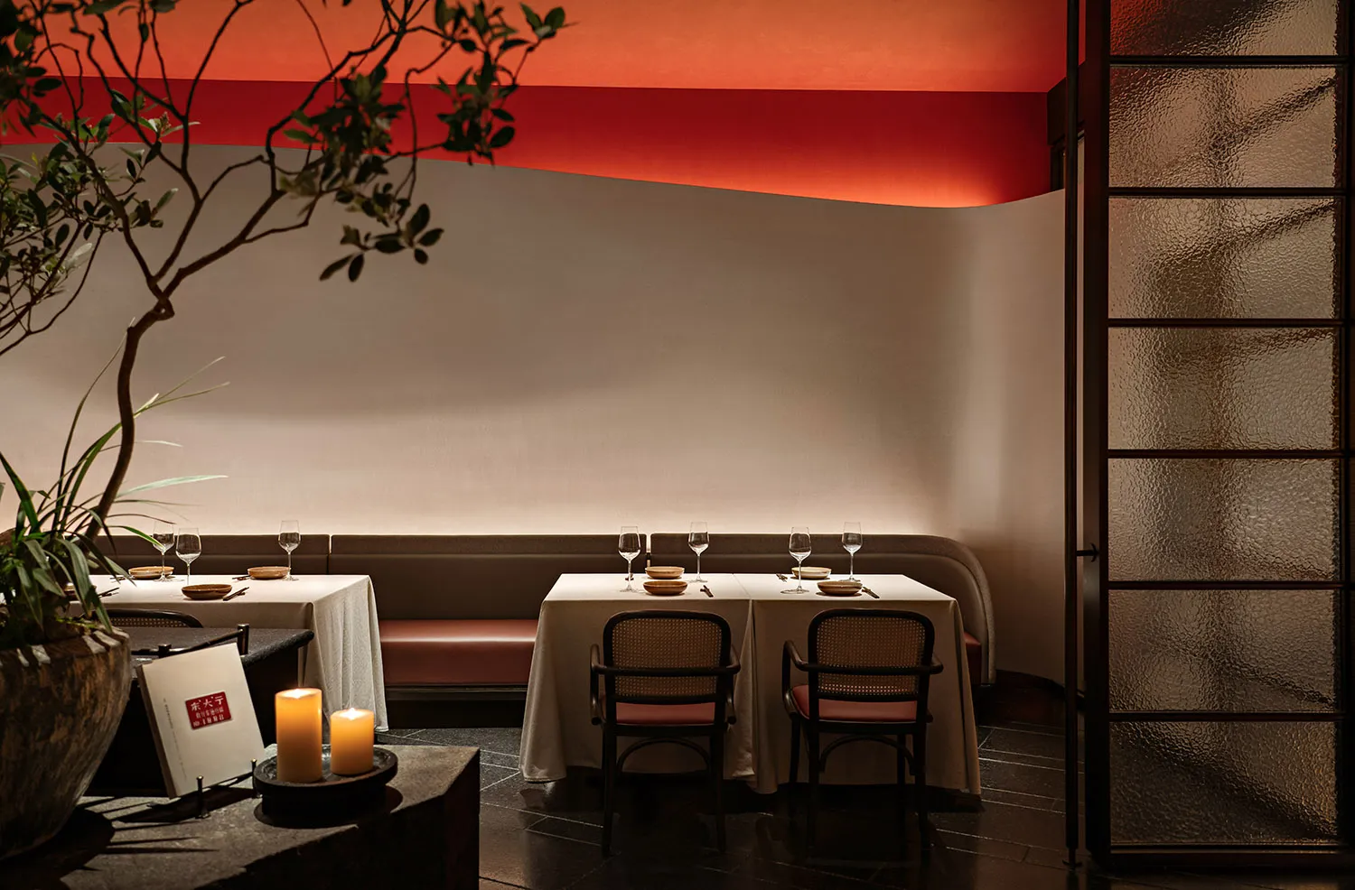 A cozy modern restaurant interior with white walls, red ceiling accents, fabric seating, candles, and potted plants.