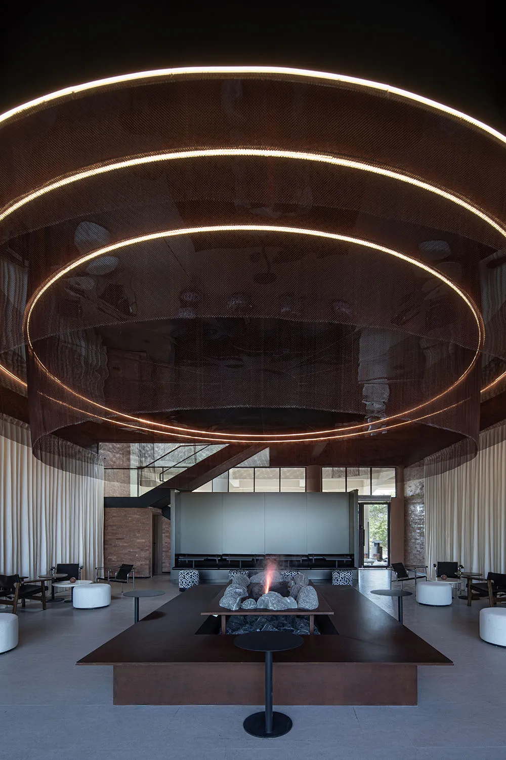 An industrial-style lobby with brick walls, metal railings, and a glass ceiling, featuring a central fire pit.
