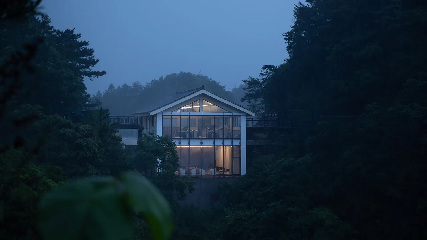 A minimalist house with a glass and wood exterior, surrounded by trees at dusk.