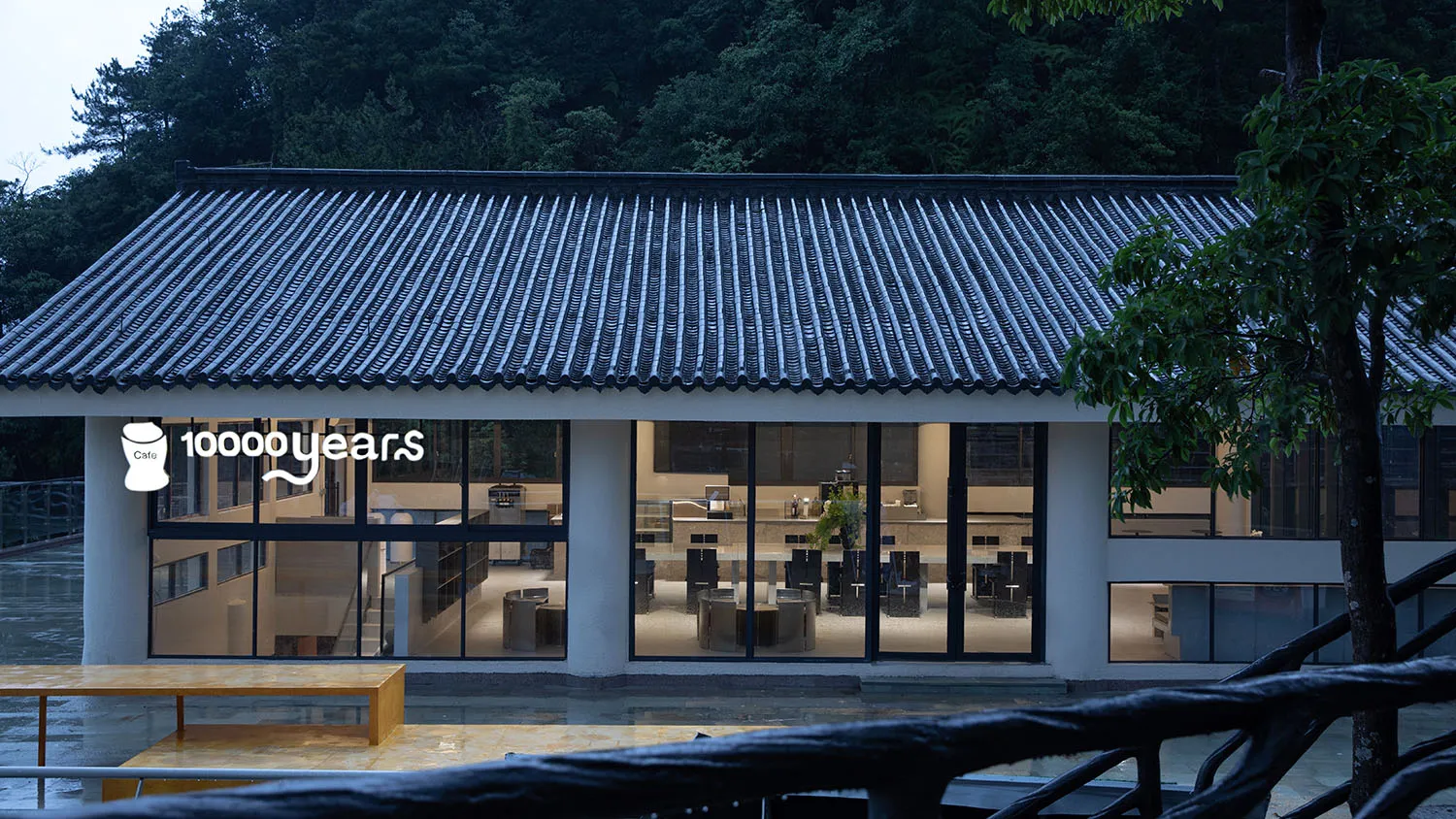 A minimalist restaurant with concrete walls and large windows, featuring modern furniture and a clean design.