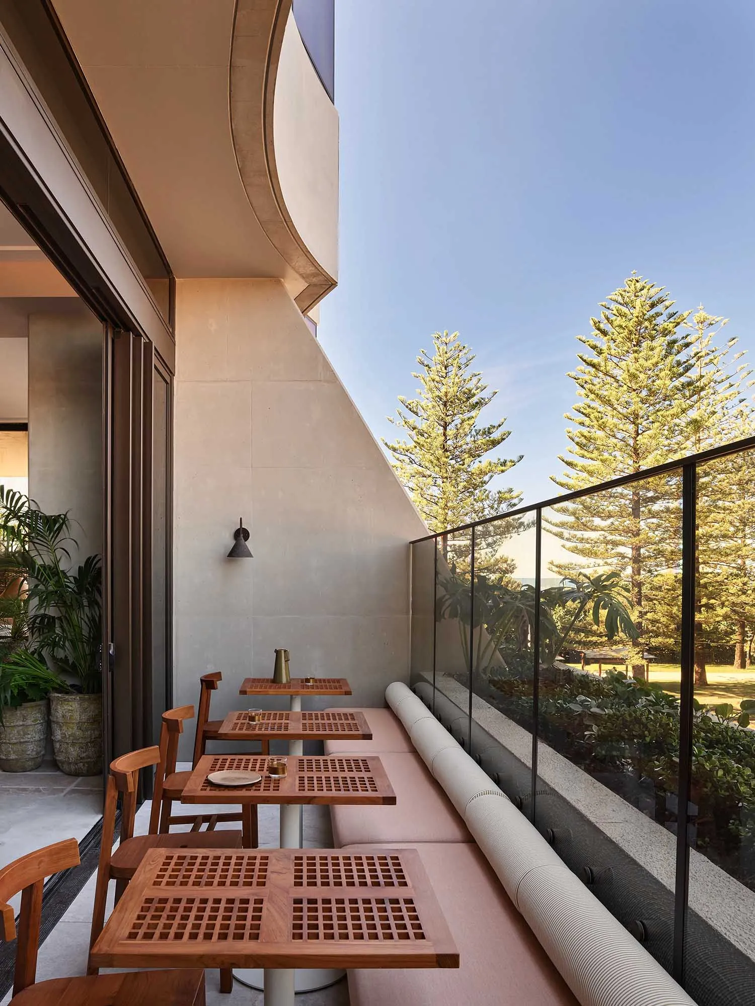 A modern restaurant balcony featuring wood furniture, concrete walls, and glass railings overlooking greenery.