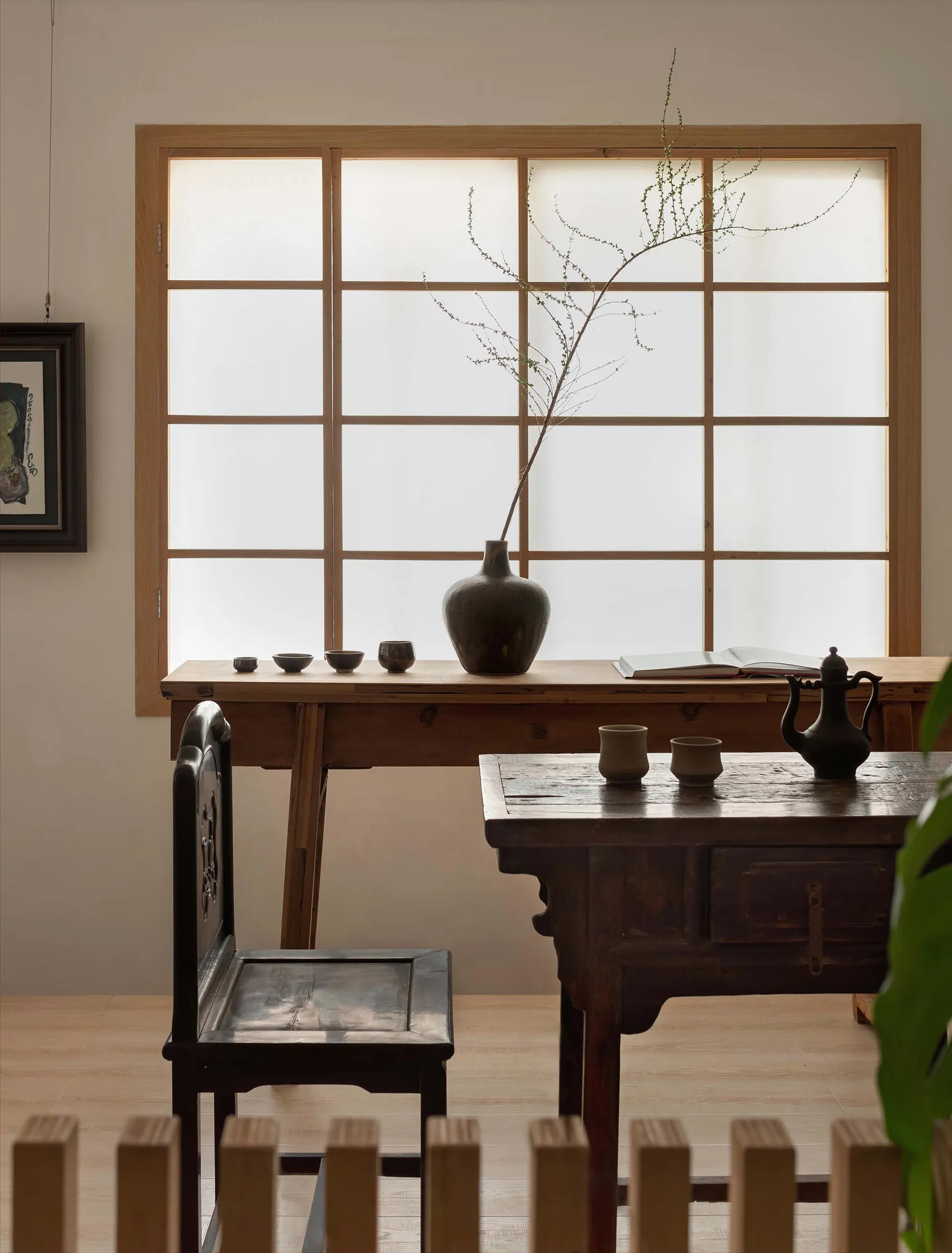 A minimalist interior featuring a wooden table, chair, and other furniture against a wall with a large window.