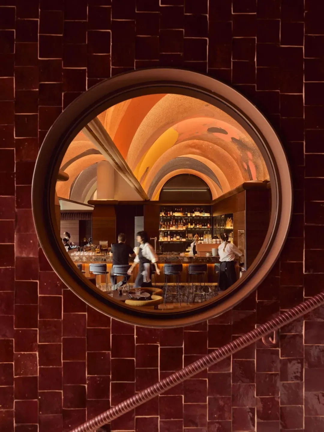 View of a restaurant bar through a circular window in a brick wall, showing a vaulted ceiling with circular arches and a well