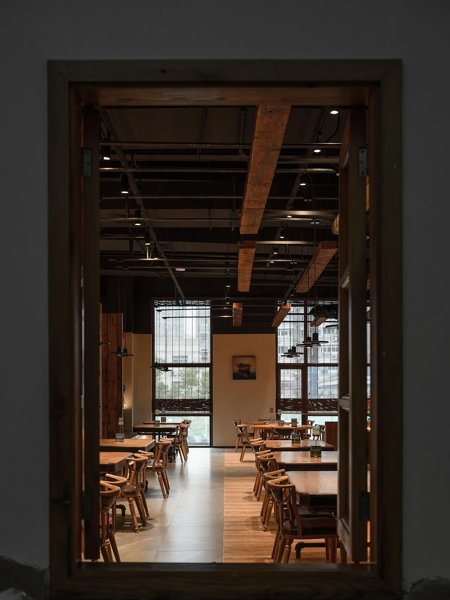 Industrial-style restaurant interior with wooden tables and chairs, large windows, and exposed beams.