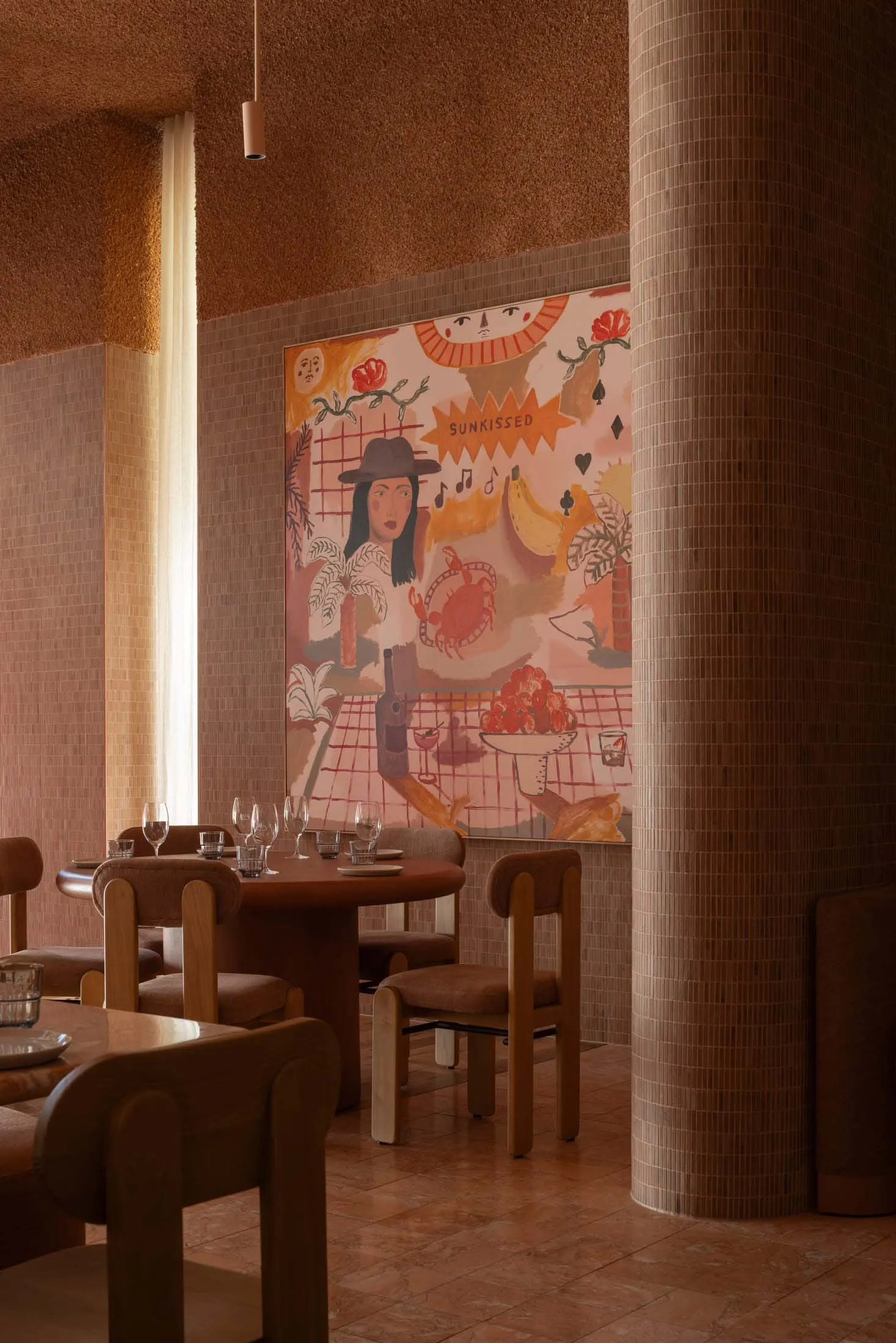 A café interior with a warm color palette, featuring chairs and tables arranged around a round table, and a colorful mural on