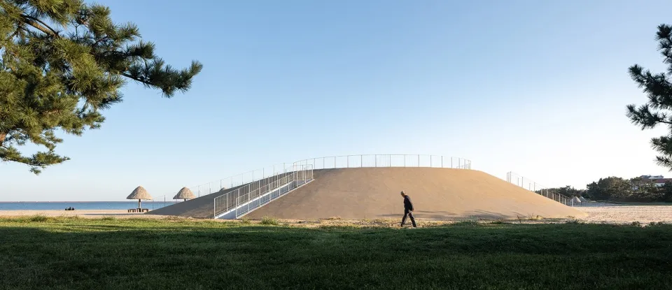 A person walking on a grassy hillside near a sandy hill with a fence on top.