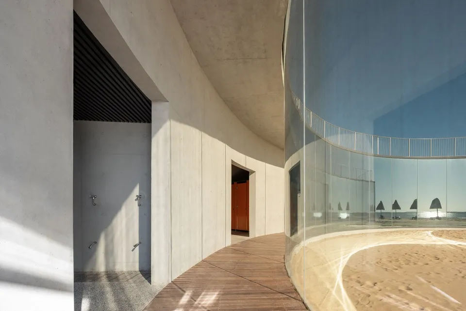 Modern beachside pavilion with concrete walls, glass walls, and wooden walkway.