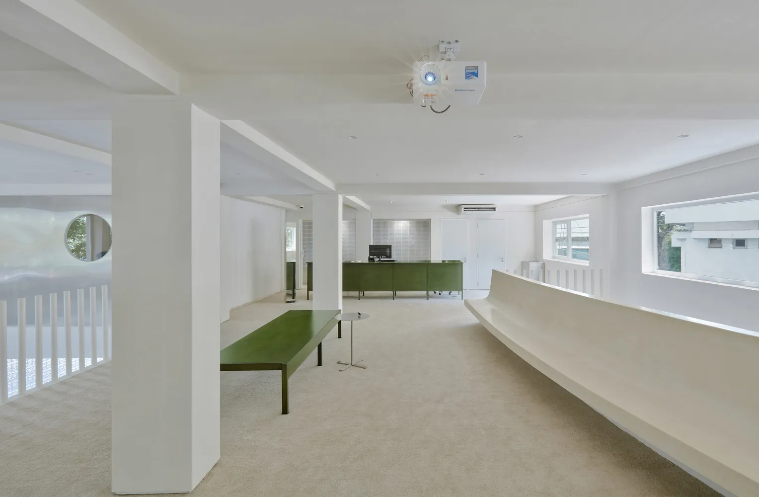 A modern office space featuring white concrete walls, a green reception desk, and minimalist furniture.
