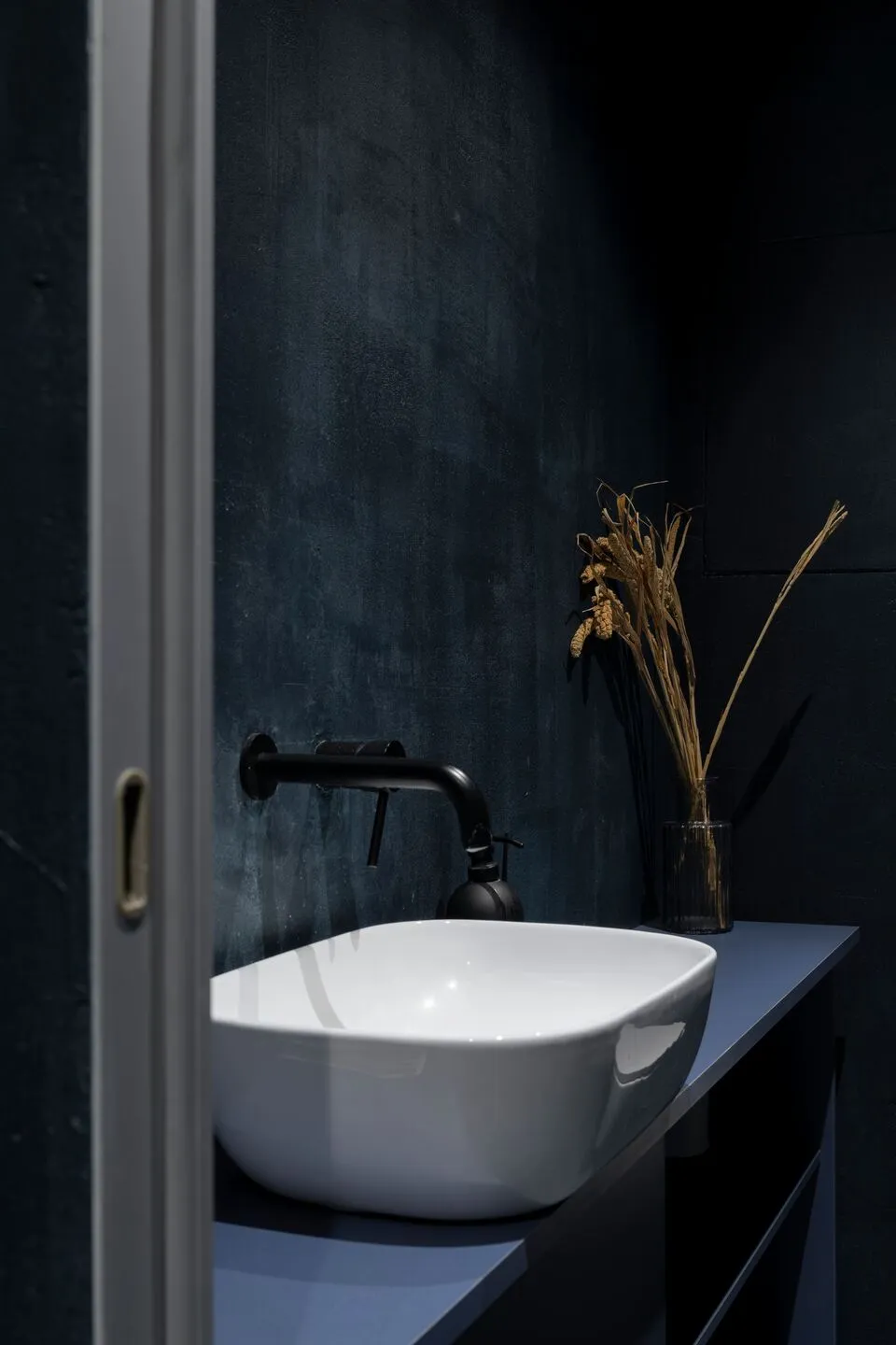 Modern minimalist bathroom with a white ceramic sink and black fixtures against a dark blue textured wall.