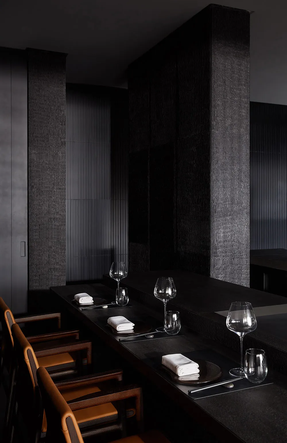 A sleek, minimalist restaurant bar with black concrete walls and counter, accompanied by elegant seating arrangements and emp