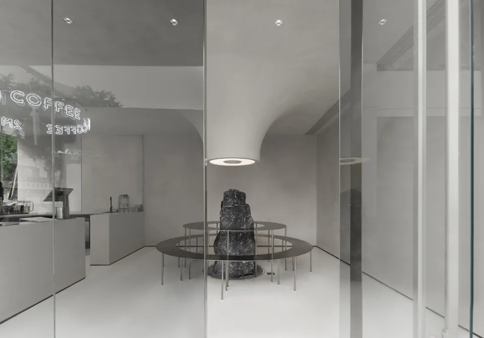 Modern coffee shop interior with minimalist design, featuring concrete surfaces and a central rock structure.