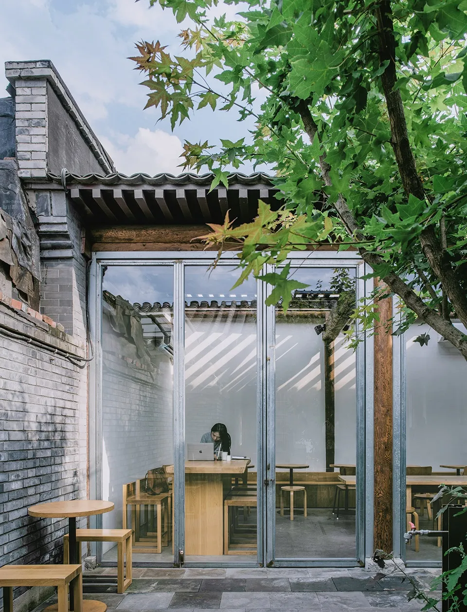 A modern coffee shop with wooden furniture and large glass doors, featuring a person working at a table inside.