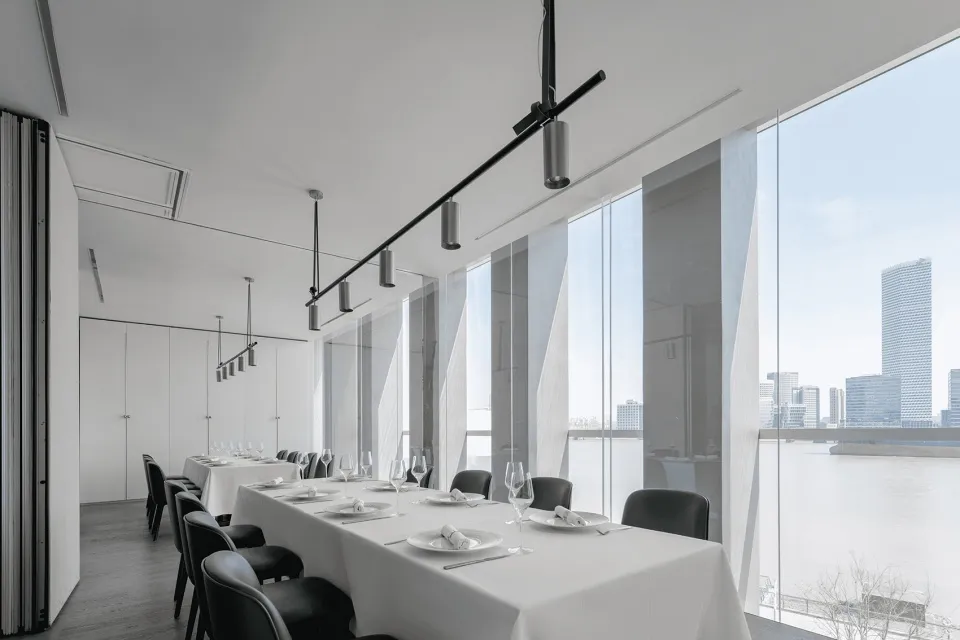 A minimalist restaurant interior with white walls and floors, featuring tables set with white tablecloths and black chairs.