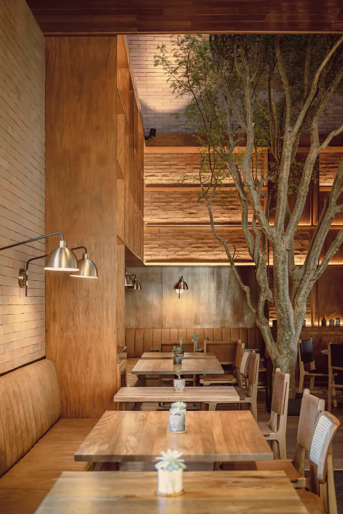 An industrial-style restaurant interior with wooden tables, wall-mounted lights, and a live tree in the center.