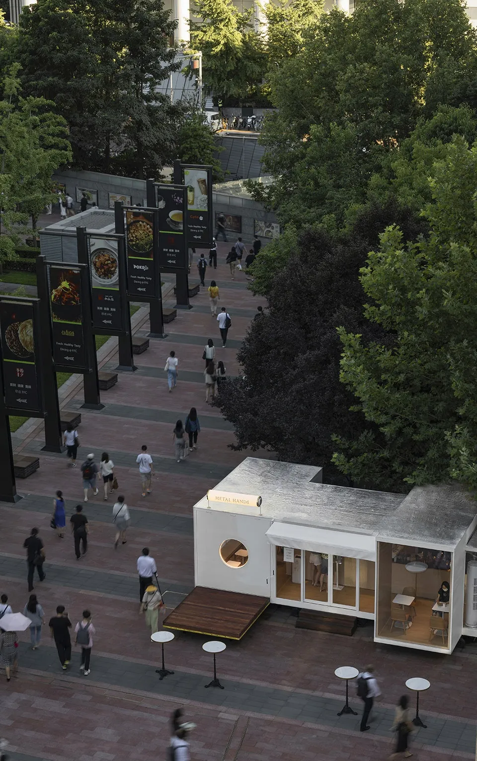 Outdoor area of a street-side cafe with metal exterior, surrounded by greenery and pedestrian walkways.