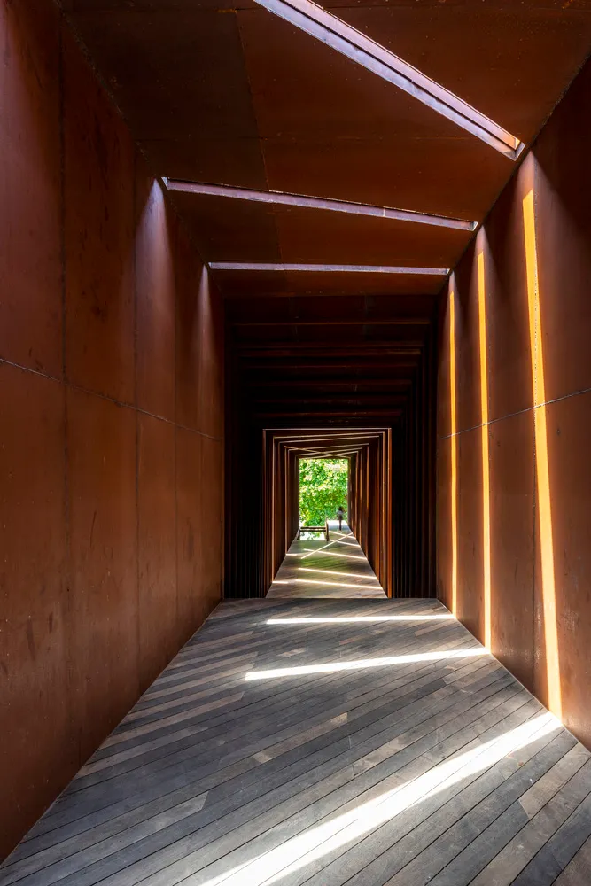 A narrow hallway lined with industrial-style wood flooring and metallic walls, leading to an open area with greenery.