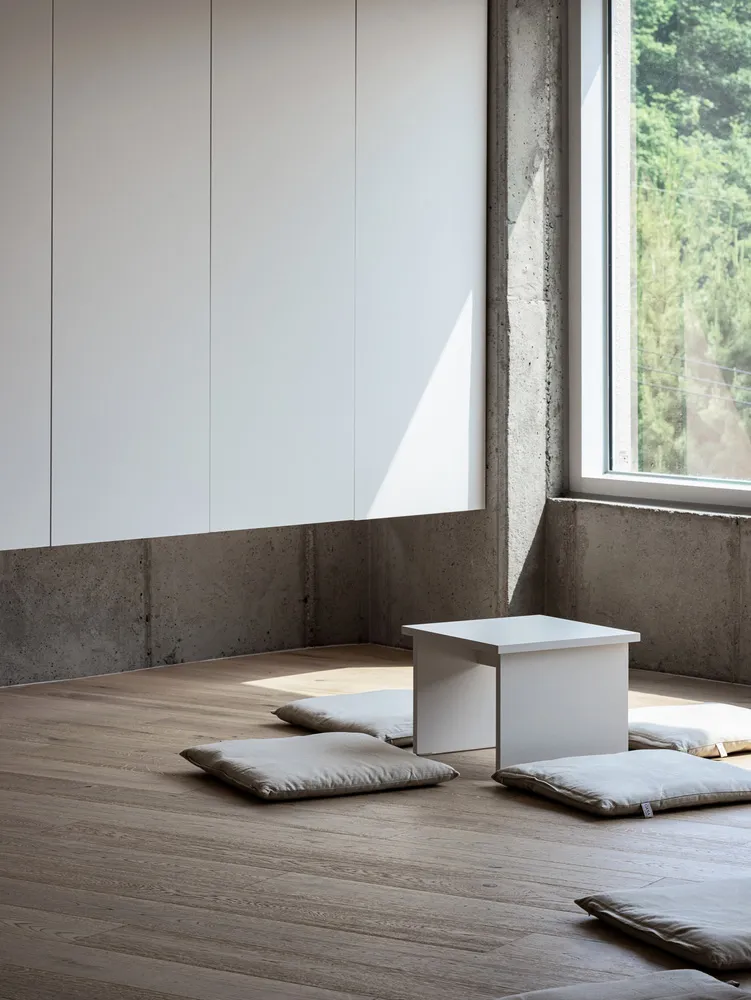 A minimalist residential space featuring concrete and wood materials, with a small white table and grey cushions on the floor