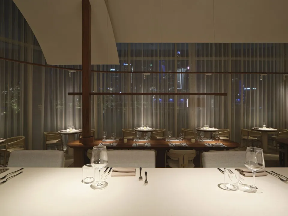 A modern dining room with glass and wooden furniture, set tables with white tablecloths and silverware, and large windows sho