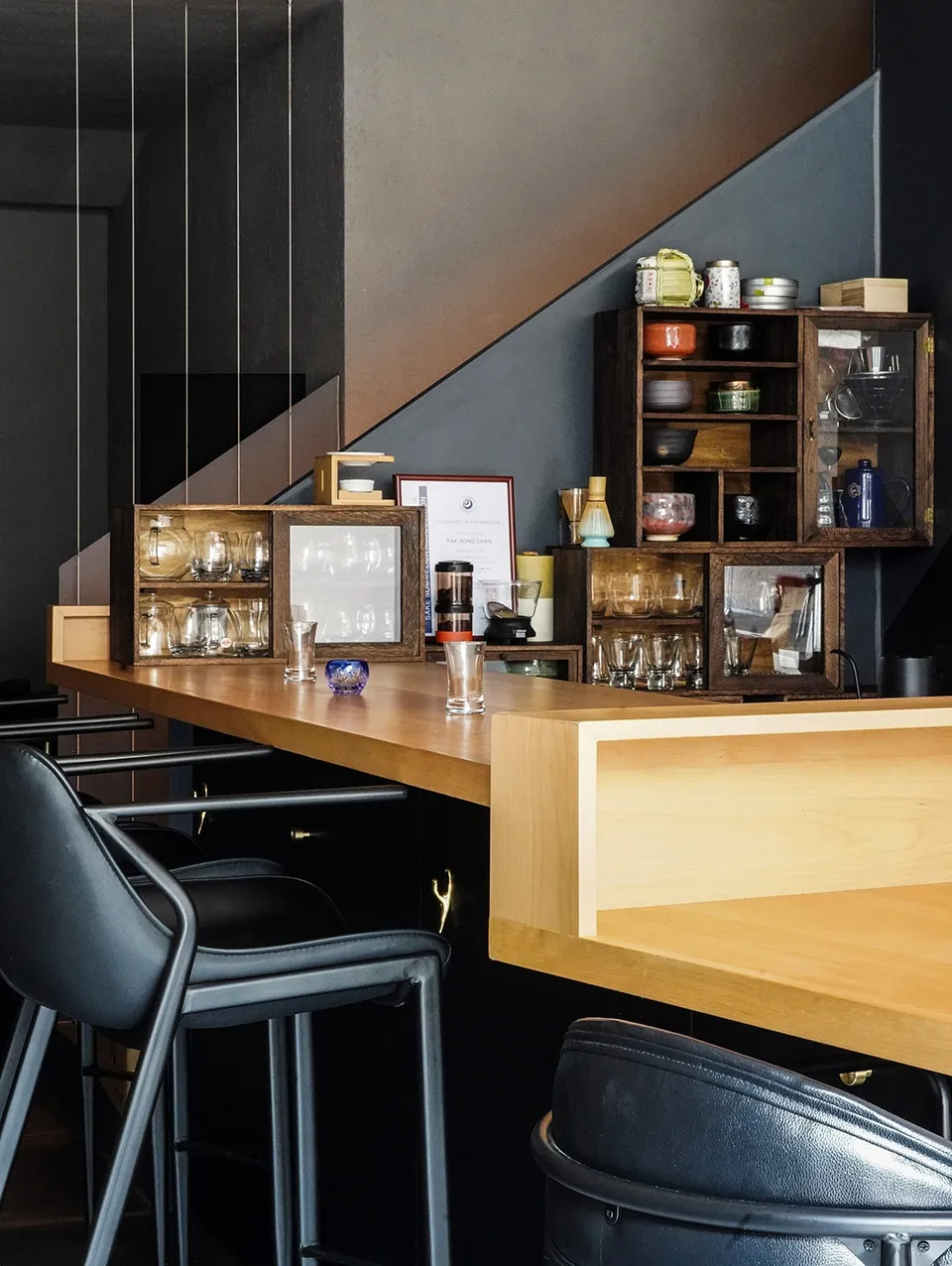 A modern restaurant bar interior featuring a wooden counter, metal stools, and various glassware and ceramic items on shelves