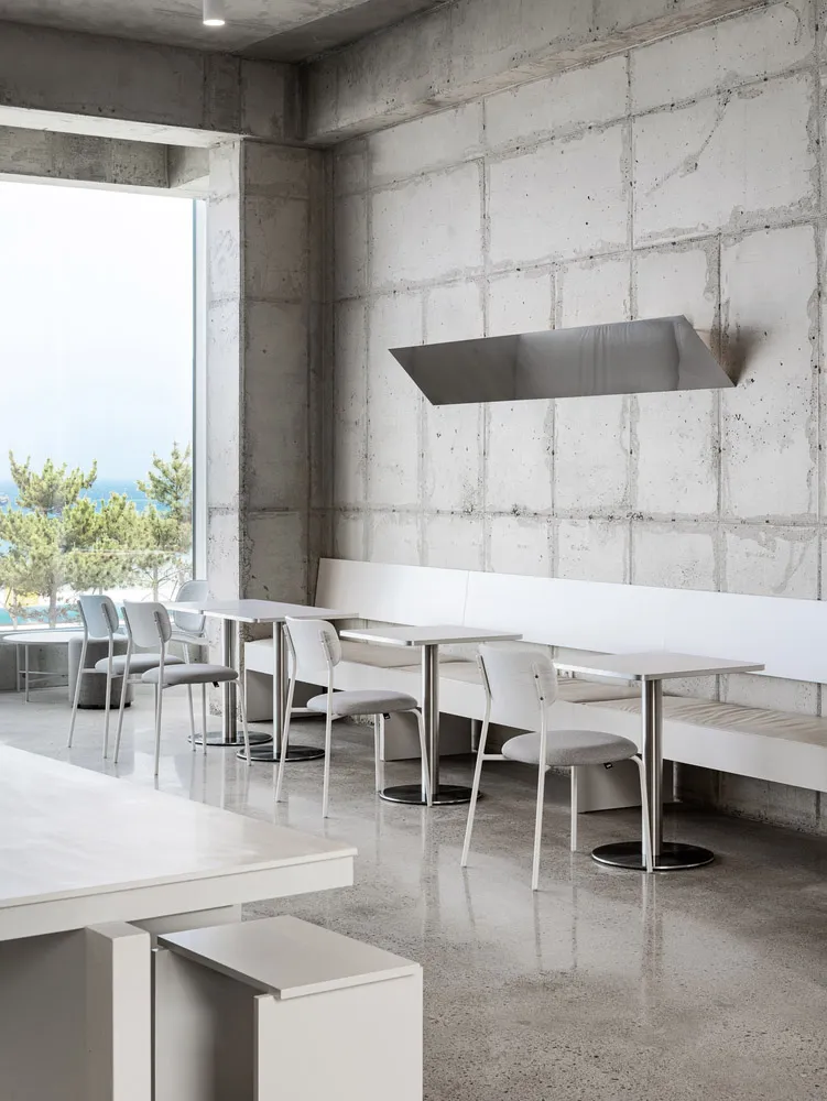 Modern restaurant interior with white tables and chairs, concrete walls, and large windows.