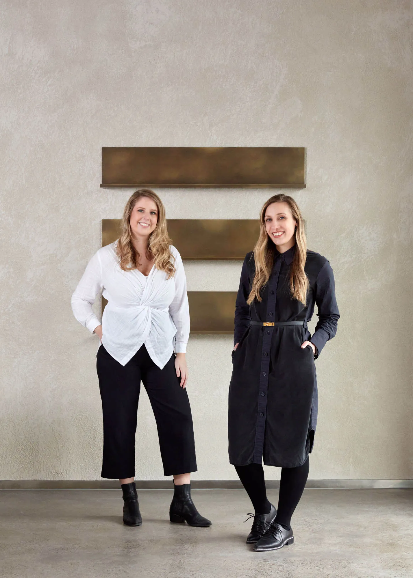 Two women wearing white and black outfits standing against a white wall with gold rectangular panels.