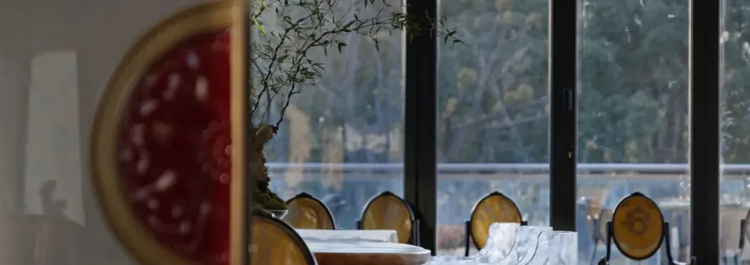 A modern restaurant interior featuring glass windows with a view of an outdoor green area and wooden tables.