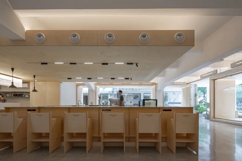 A minimalist restaurant/bar interior with wooden tables and chairs, open kitchen, and large windows.