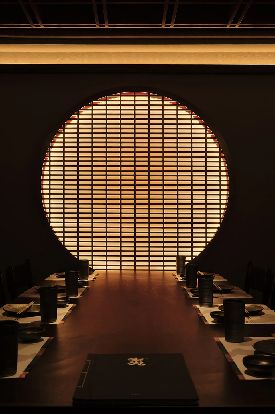 A long dark restaurant dining table with plates, chopsticks, and cups set for guests, with a large circular window in the bac