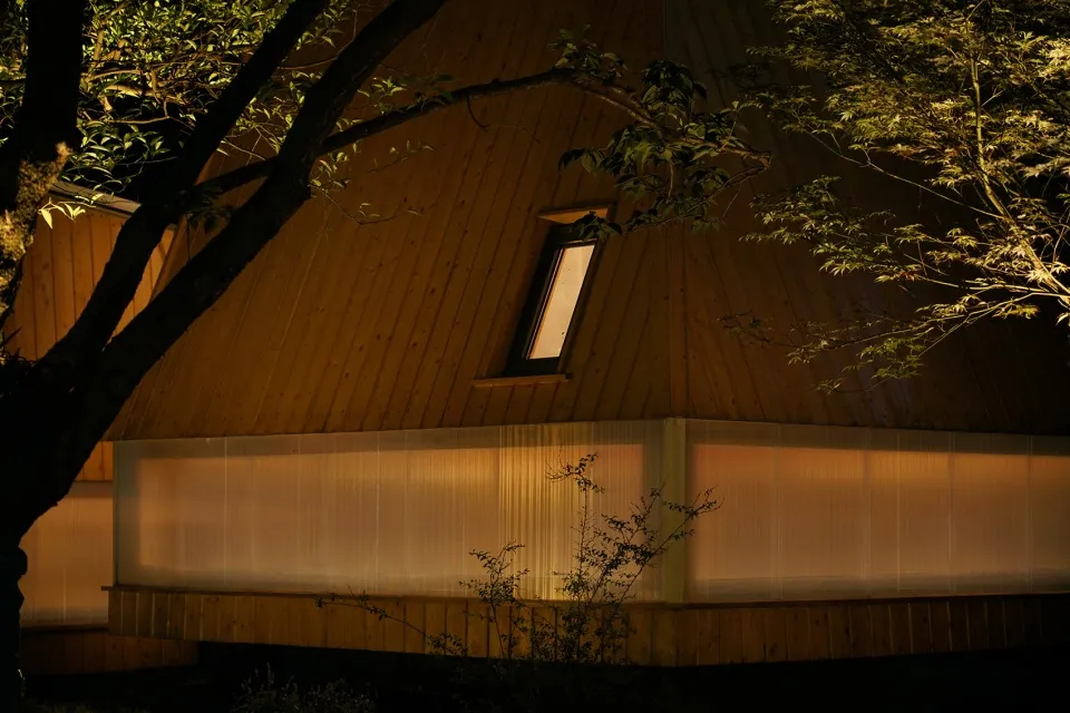 A modern house at night, featuring wooden exterior and a single illuminated window.