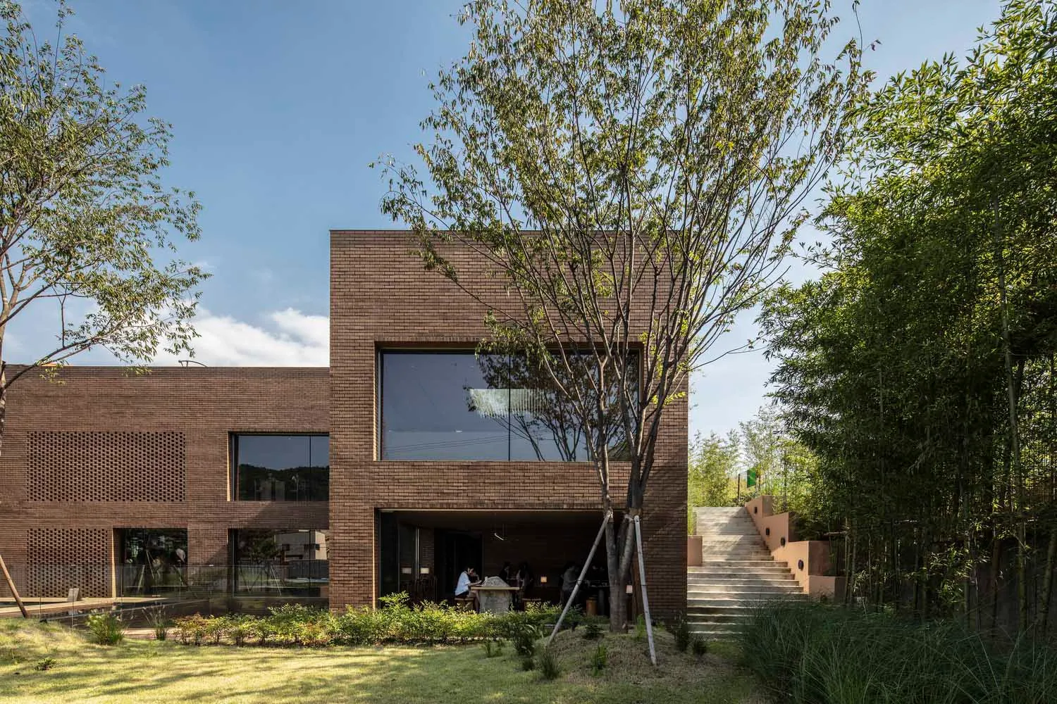 A modern, brick building with large windows surrounded by greenery and steps.