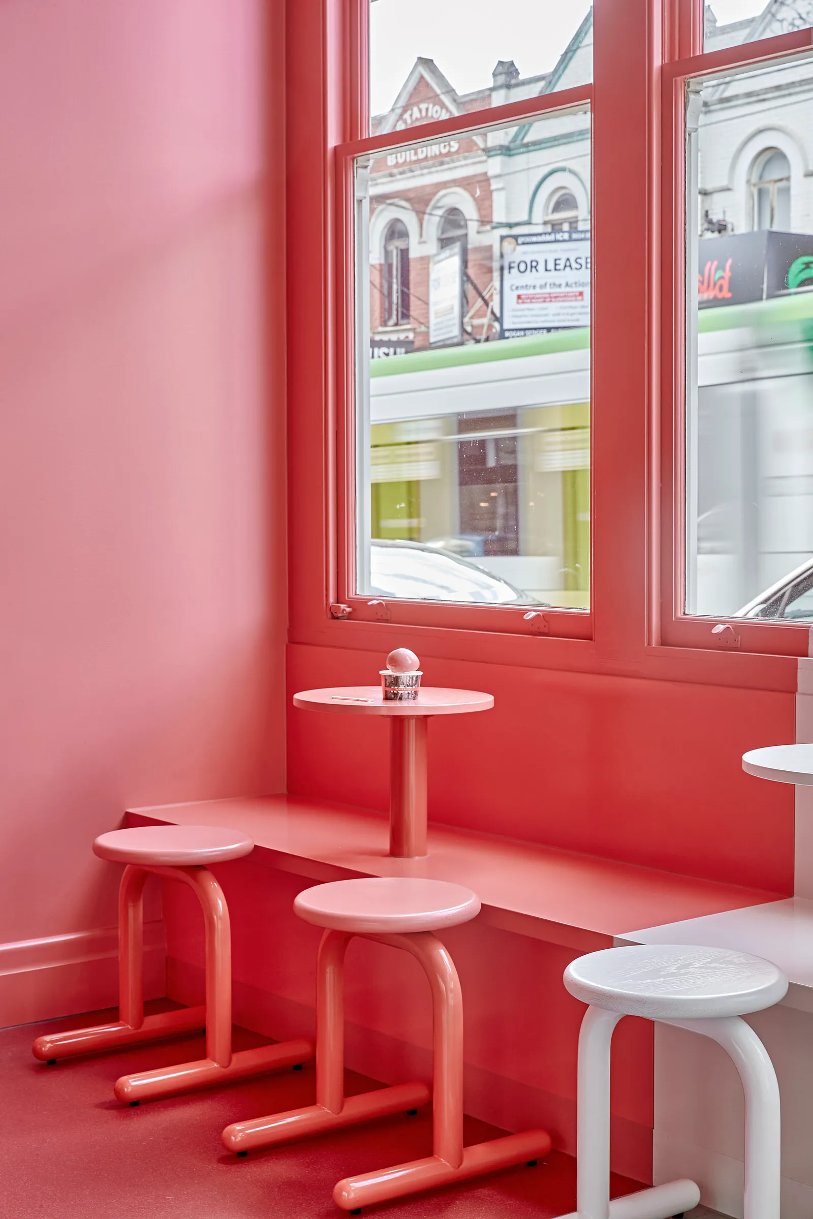 A pink, minimalist restaurant interior with a pink table and stools, and a window overlooking buildings.