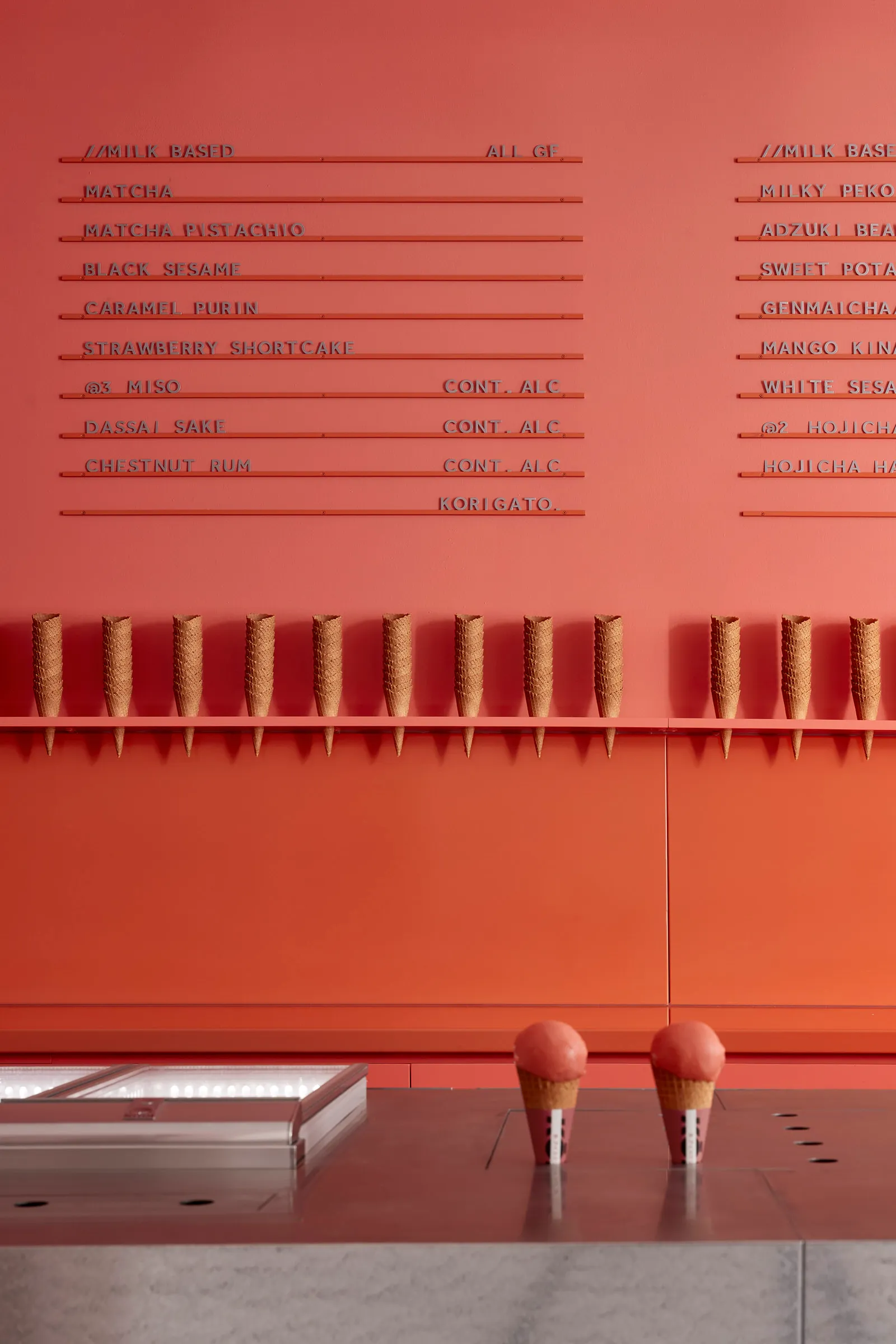 A modern, minimalist food court interior with concrete walls displaying an extensive menu of ice cream flavors and cones.